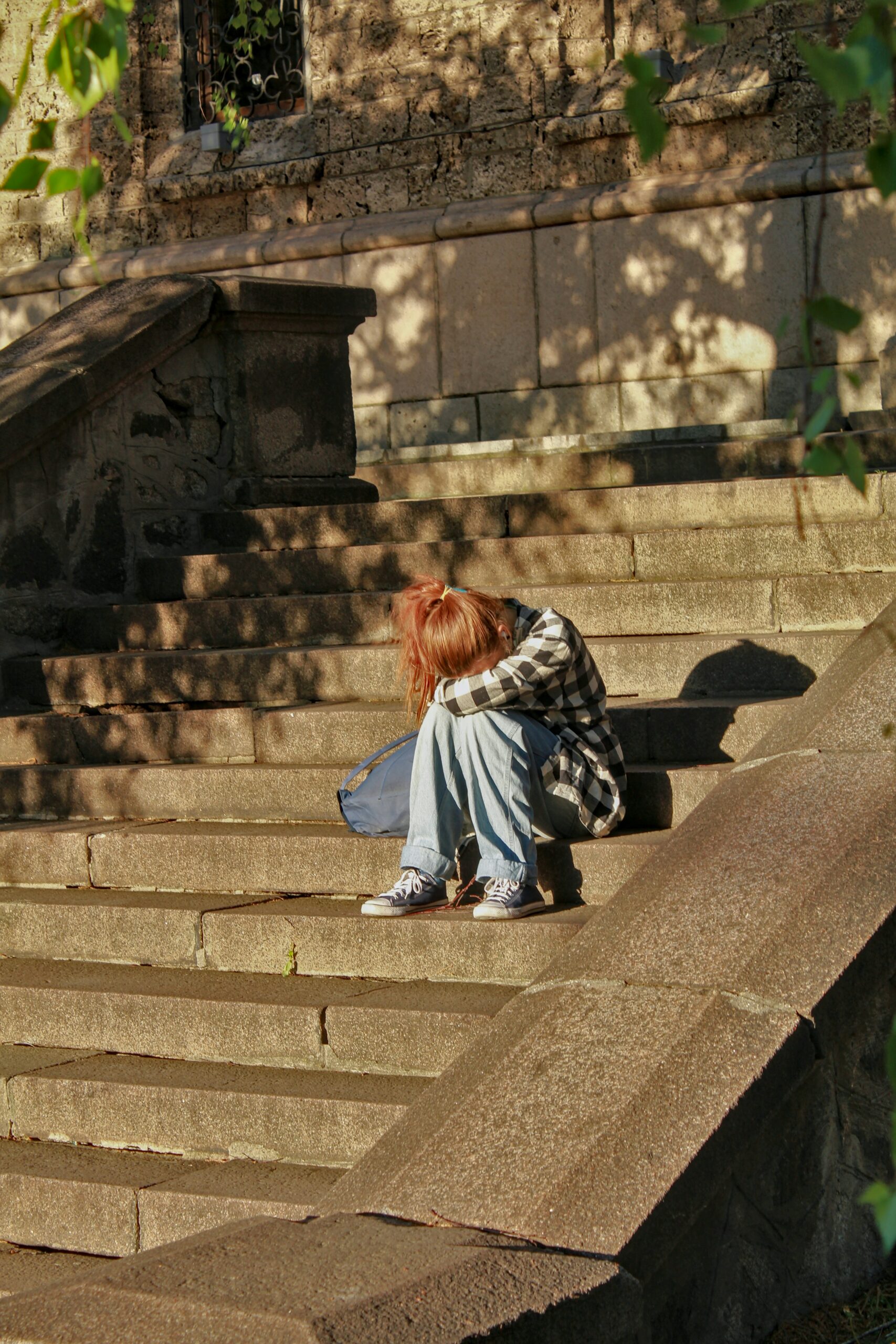 A teenage girl, symbolising the emotional impact of child emancipation laws in Australia, sits on the steps, her head bowed forward resting on her knees, in a contemplative and possibly troubled pose.