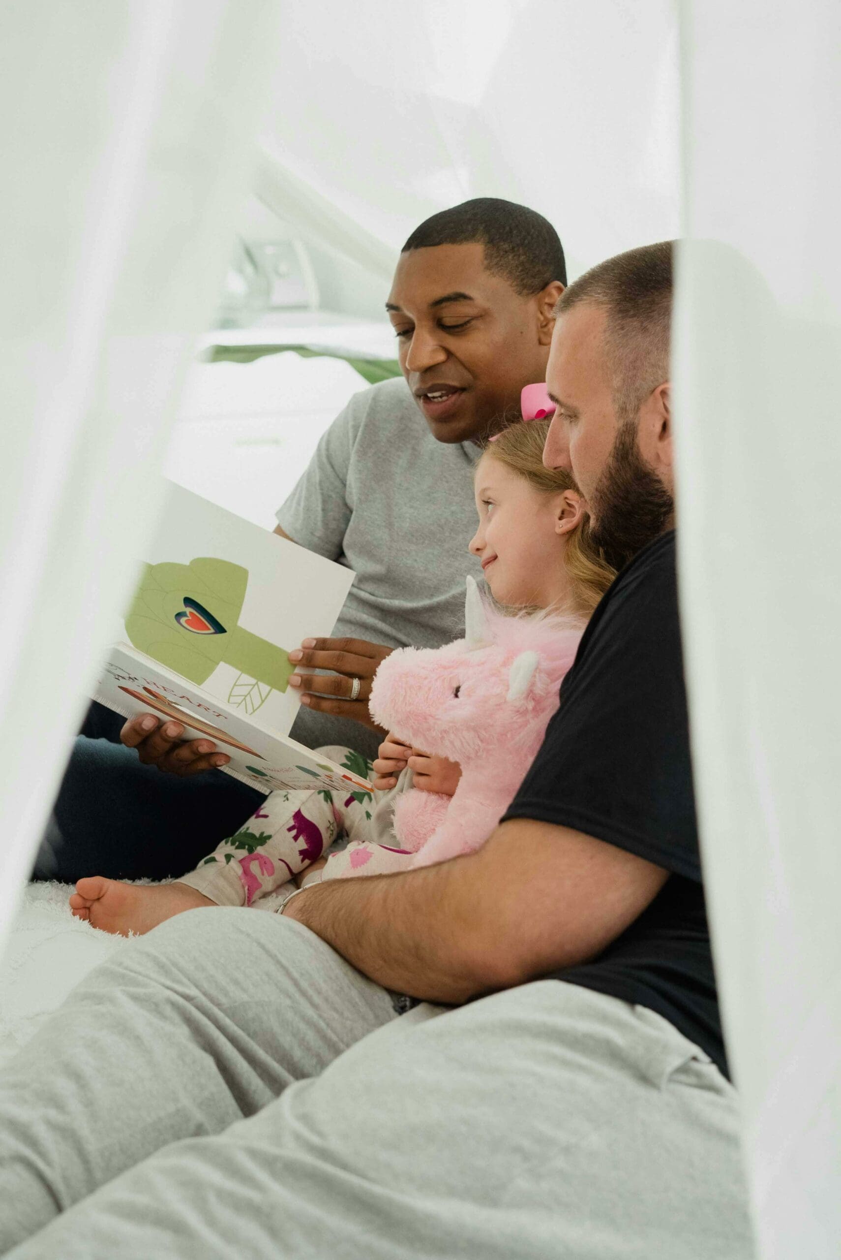 A joyful moment inside a cozy blanket fort with two men and a young child reading a book together, illustrating the nurturing environment created through the adoption process in Australia.